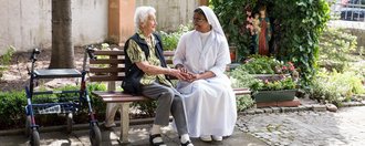 im Garten auf einer Bank: eine Ordensschwester hält die Hand einer Bewohnerin