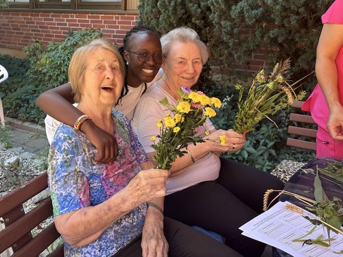 Drei Frauen, zwei ältere Damen und eine jüngere Frau, sitzen eng beieinander auf einer Bank im Freien. Alle halten kleine Blumensträuße in den Händen und strahlen vor Freude. Die jüngere Frau hat ihren Arm liebevoll um eine der Seniorinnen gelegt.