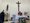 Ein Priester in roten Gewändern steht hinter einem Altar und feiert die Messe. An der weißen Wand dahinter hängt ein Kruzifix. Links steht eine Person in schwarzer Kleidung und hält Palmzweige.
