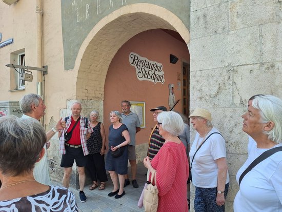 Eine Gruppe älterer Menschen steht vor einem steinernen Torbogen mit der Aufschrift „Restaurant St. Georg“. Die Personen blicken aufmerksam in eine Richtung, als würden sie einer Führung oder Ansprache lauschen.