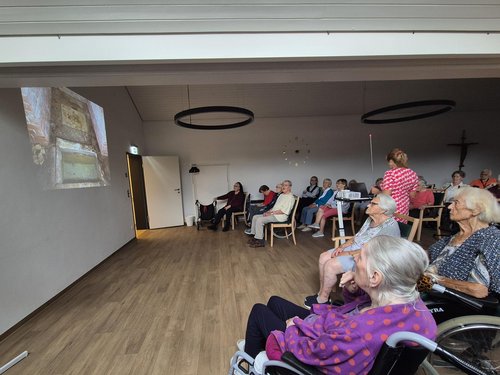 Eine Gruppe älterer Menschen, teilweise im Rollstuhl, sitzt in einem hellen Raum und schaut gemeinsam auf eine Bildprojektion an der Wand.
