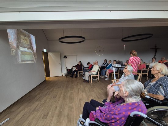 Eine Gruppe älterer Menschen, teilweise im Rollstuhl, sitzt in einem hellen Raum und schaut gemeinsam auf eine Bildprojektion an der Wand.