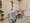 Ein Priester in weißen Gewändern steht an einem steinernen Lesepult in einer Kirche und liest vor. Im Hintergrund steht ein weiterer Mann am Altar unter einem großen Kreuz an der Wand. Blumen schmücken den Altarraum.