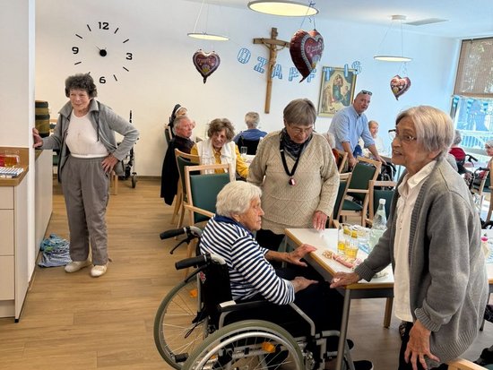 Ältere Menschen in einem hellen Gemeinschaftsraum. Eine Frau im Rollstuhl sitzt an einem Tisch und unterhält sich mit einer stehenden Dame. Im Hintergrund sind weitere Senioren zu sehen. An der Wand hängen ein großes Zifferblatt und Herz-Ballons.