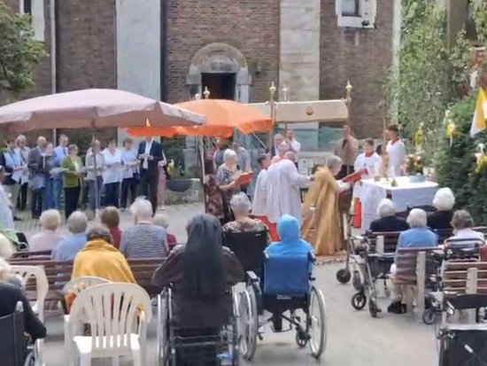 Ältere Menschen, teilweise im Rollstuhl, sitzen im Freien vor einer Kirche und nehmen an einem Gottesdienst teil. Priester in Gewändern stehen am Altar, während ein Chor im Hintergrund unter Sonnenschirmen singt.