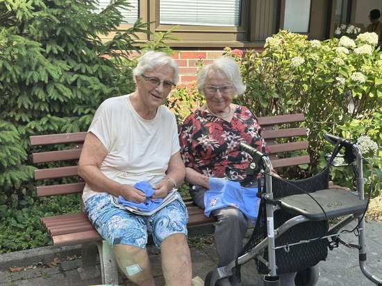 Zwei lächelnde Seniorinnen sitzen auf einer Parkbank im Freien. Rechts neben der Bank steht ein Rollator. Beide halten blaue Stoffe in den Händen.