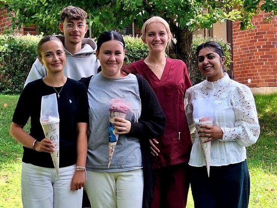 Fünf junge Erwachsene stehen lächelnd im Freien auf einer Wiese und halten gerollte Urkunden oder Diplome in den Händen. Die Szene deutet auf eine Abschlussfeier oder eine feierliche Übergabe von Zertifikaten hin. Im Hintergrund sind grüne Bäume zu sehen.