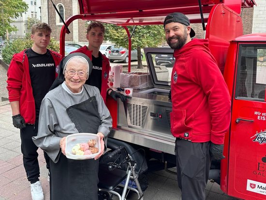 Eine ältere Nonne mit Gehhilfe hält einen Teller mit Essen und steht vor einem roten Foodtruck. Drei Männer in roten Jacken stehen lächelnd neben ihr. Im Hintergrund ist eine Kirche zu sehen.
