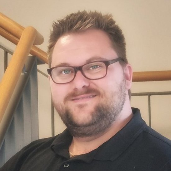 Ein Mann mit Brille, Bart und schwarzem Poloshirt lächelt in die Kamera. Im Hintergrund ist ein Treppengeländer zu sehen.