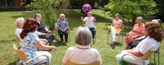 Bewohner/innen, Pflegerinnen und Ordensschwestern sitzen im Garten auf Stühlen im Kreis und werfen sich einen Ball zu