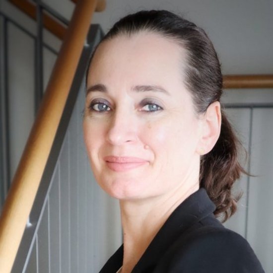 Eine Frau mit zurückgebundenen dunklen Haaren und einem schwarzen Blazer steht lächelnd an einer Treppe. Sie hält das hölzerne Handlauf und trägt eine Uhr am Handgelenk.